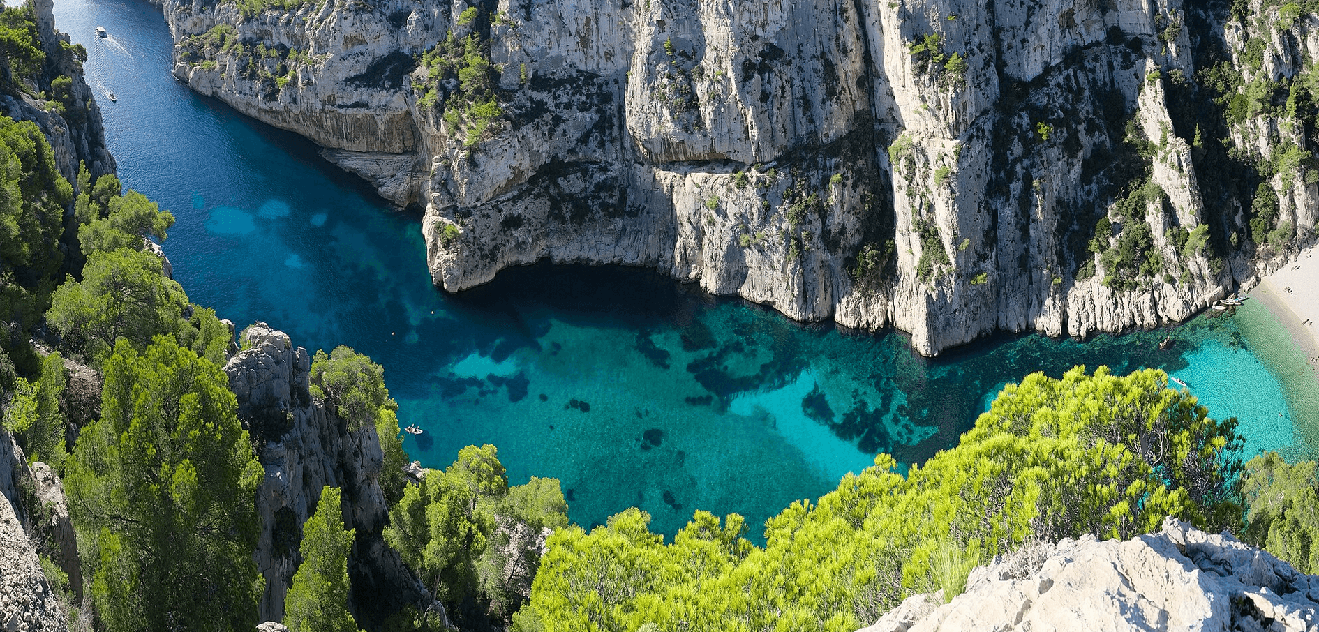 Vue plongeante sur la calanque d’En-Vau, entourée de hautes falaises calcaires et de pins méditerranéens, avec une mer turquoise et une plage de galets en contrebas.