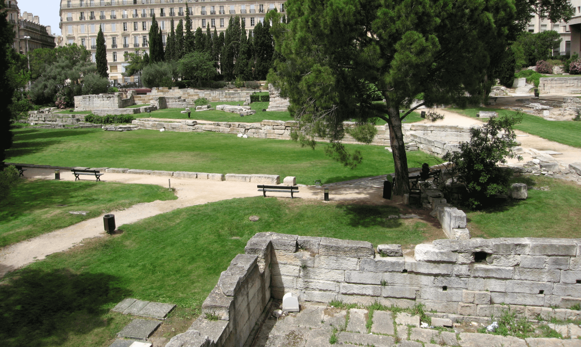 Vue sur le Jardin des Vestiges à Marseille, avec des ruines antiques en pierre, des pelouses bien entretenues, des bancs et un chemin de promenade, en arrière-plan un immeuble haussmannien.