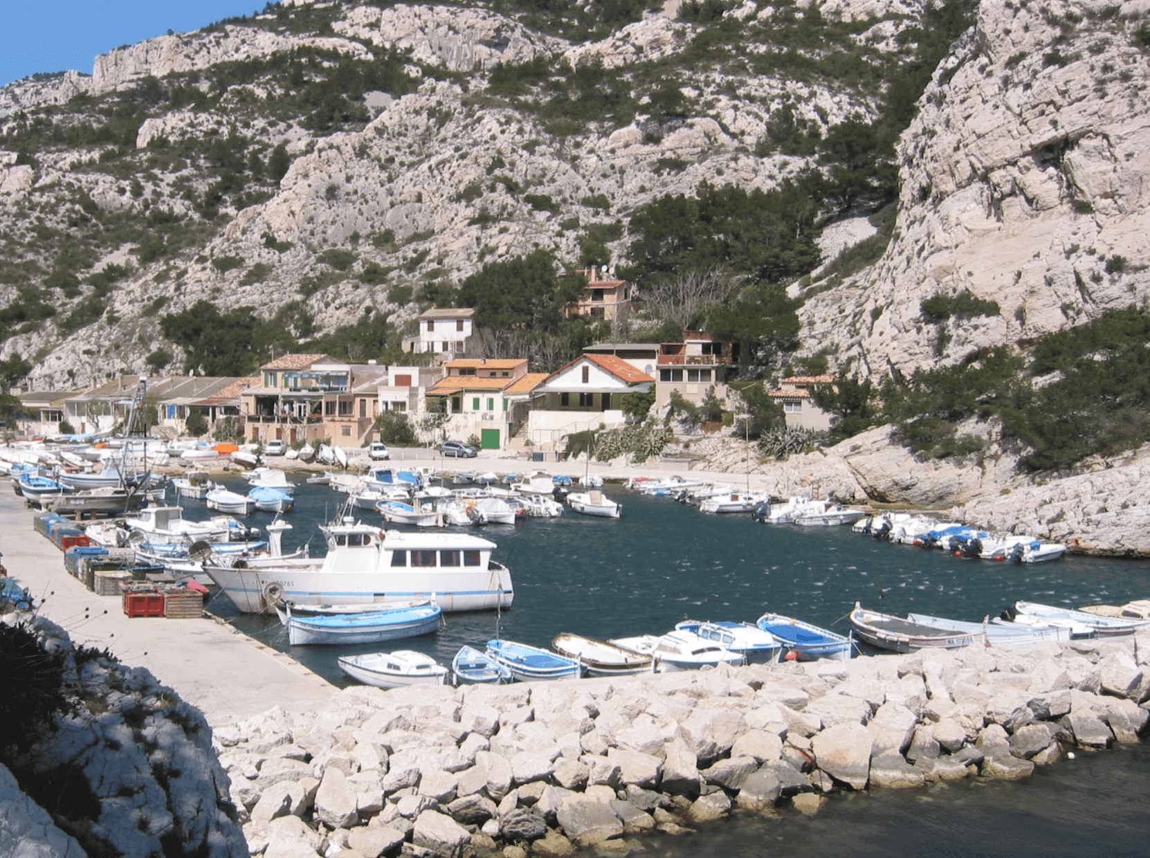 Petit port pittoresque de la calanque de Morgiou, avec ses barques amarrées, ses maisons traditionnelles nichées au pied des falaises calcaires et une mer turquoise bordée de végétation méditerranéenne.