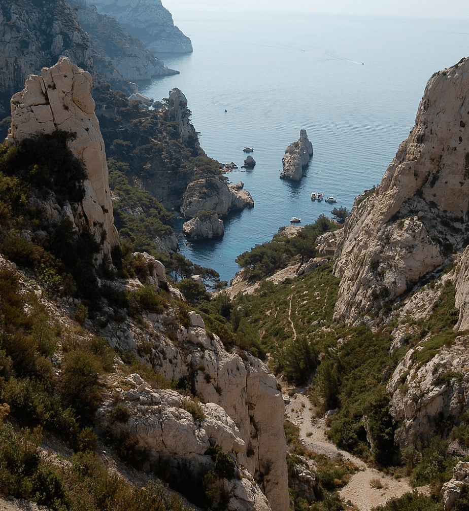 Vue plongeante sur la calanque de Sugiton, encadrée par des falaises escarpées et parsemée de rochers dans une mer d’un bleu profond, avec des sentiers sinueux bordés de végétation méditerranéenne.