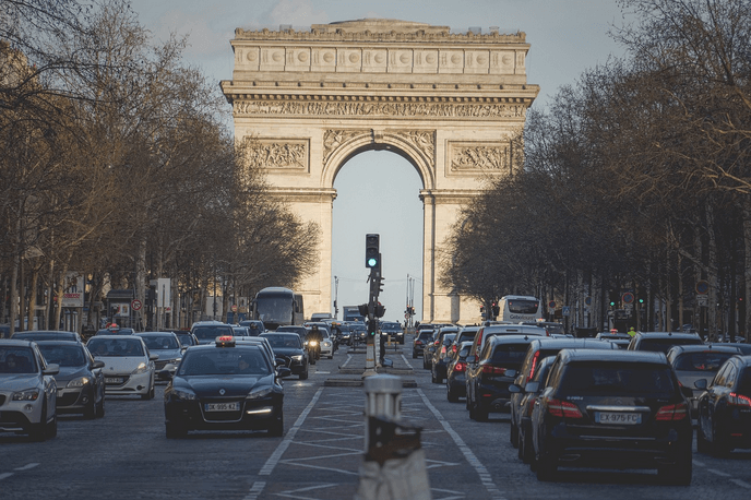 Vue de l’avenue des Champs-Élysées bordée d’arbres, avec des voitures circulant au centre. À l’arrière-plan, l’Arc de Triomphe est visible.