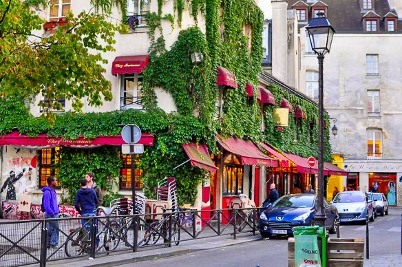 Ruelle pavée du quartier du Marais à Paris, bordée de bâtiments anciens aux façades claires, de pots de fleurs turquoise et d’un lampadaire central. Le ciel est couvert, donnant une atmosphère calme et légèrement mélancolique.