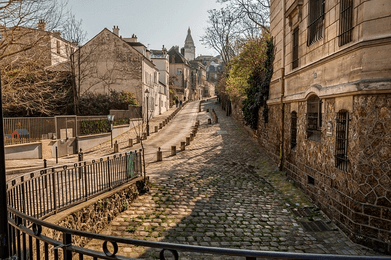 Rue pavée en pente du quartier de Montmartre à Paris, bordée de bâtiments anciens et de murets en pierre, avec la basilique du Sacré-Cœur visible au loin sous un ciel clair.