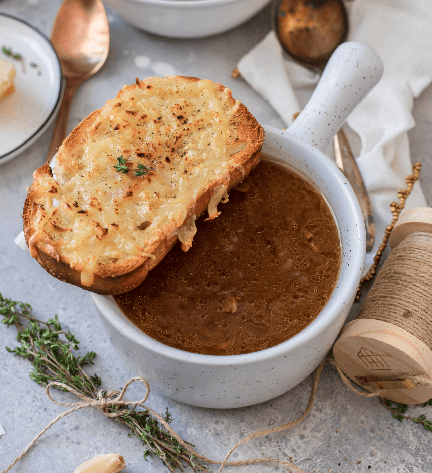 Soupe à l’oignon servie dans un bol blanc, accompagnée d’un pain grillé au fromage gratiné posé sur le rebord.